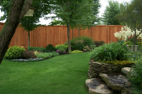 Manicured lawn bordered by lush garden beds, a stacked stone wall, and a tall wood privacy fence.