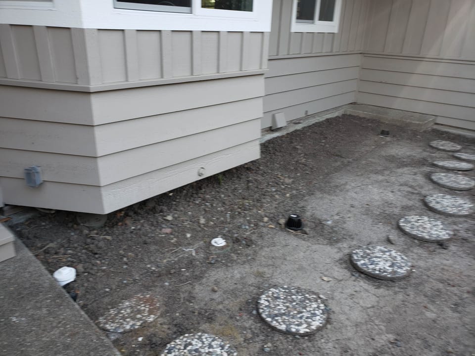 Bare dirt ground with stepping stones and pipe cleanout caps next to a house corner.