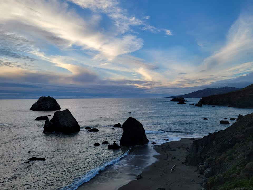 Rocky sea stacks sit in ocean waters along a curved sandy beach and coastal cliffs under an evening sky.