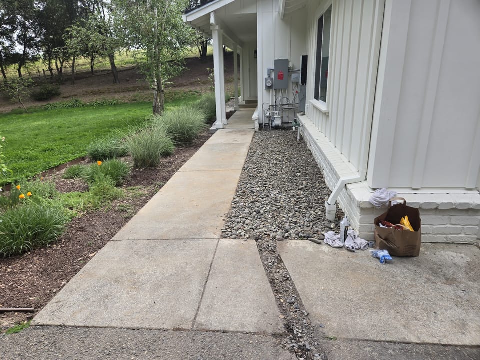 A rock gravel border and concrete sidewalk run along the side of a white house next to a lawn.