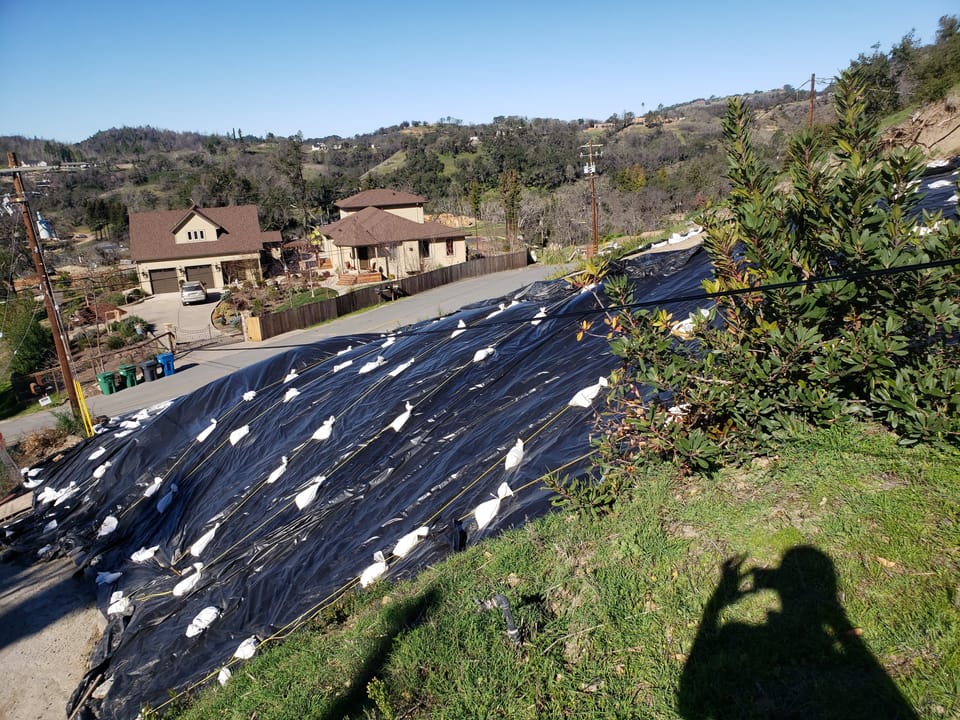 Black plastic sheeting held down by sandbags covers a steep residential hillside to prevent erosion above a road.