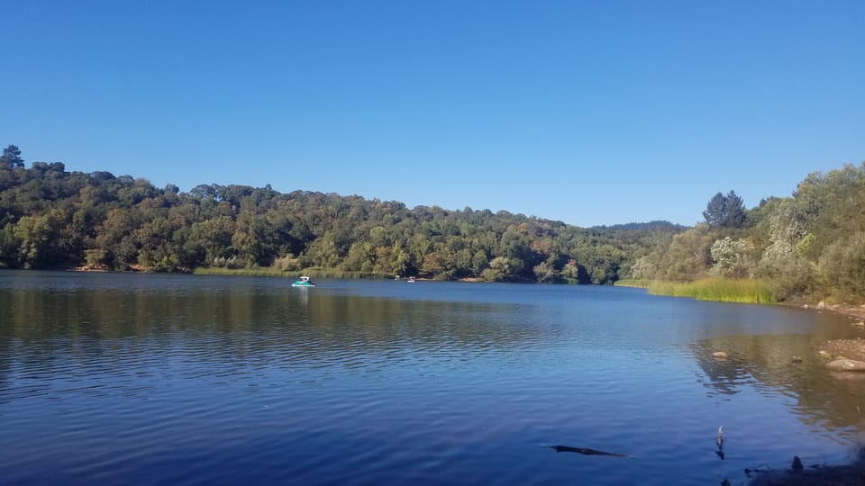 Wide calm blue lake surrounded by forested hills under a clear sky, with small paddle boats in the distance.