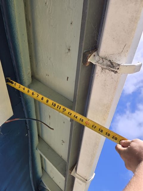A hand holds a yellow tape measure under an exterior house eave next to a white downspout.