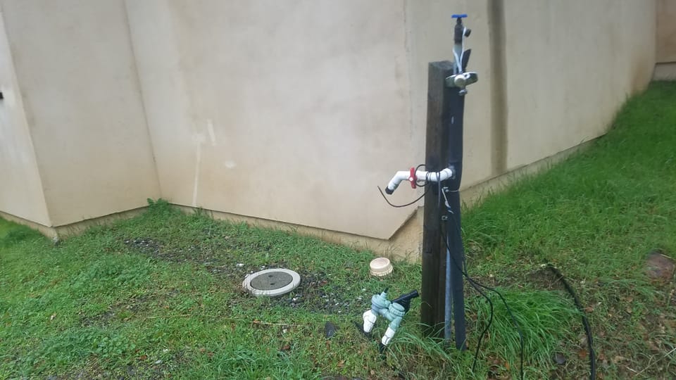 Plumbing pipes, spigots, and shutoff valves mounted to a wooden post beside a stucco building foundation.
