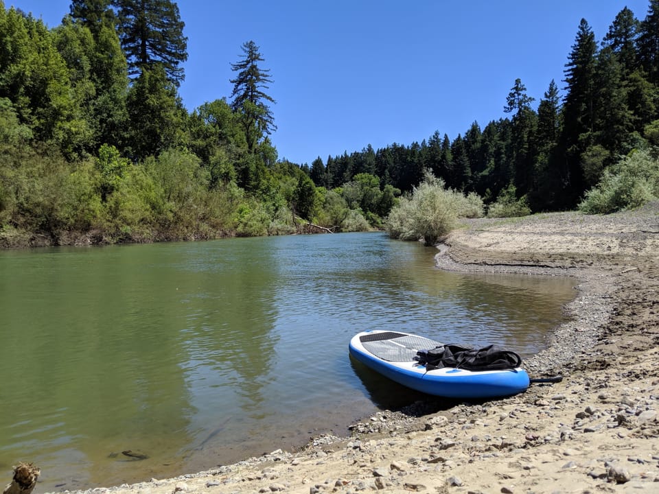 Inflatable paddleboard rests on a gravel riverbank beside calm green water bordered by tall forest.