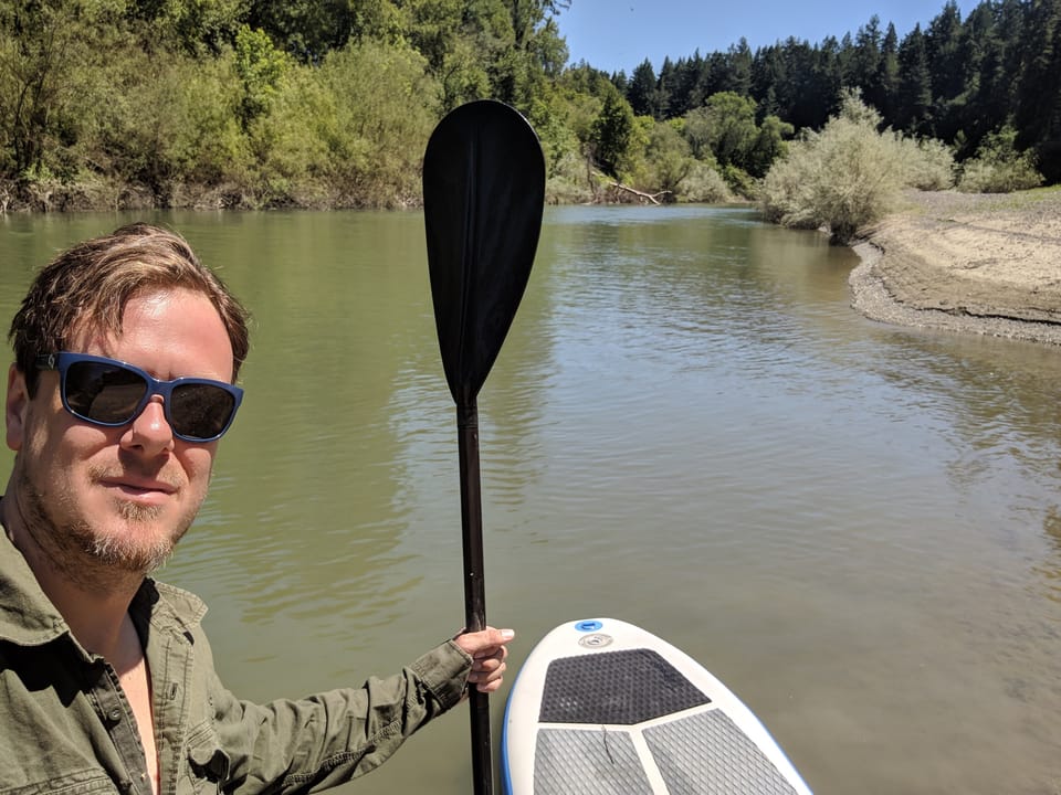Devan wearing sunglasses holds a paddle on a stand-up paddleboard floating on a calm river with wooded banks.