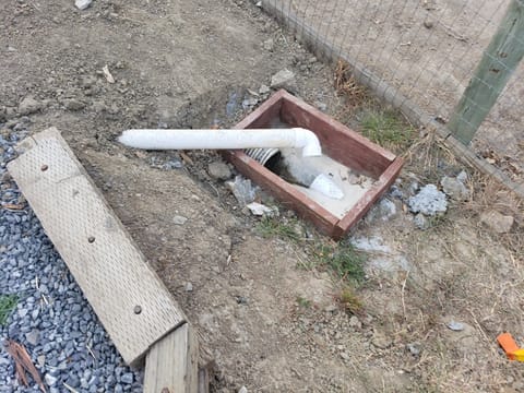 White PVC pipe discharging into a small wooden framed ground basin next to a wire fence.