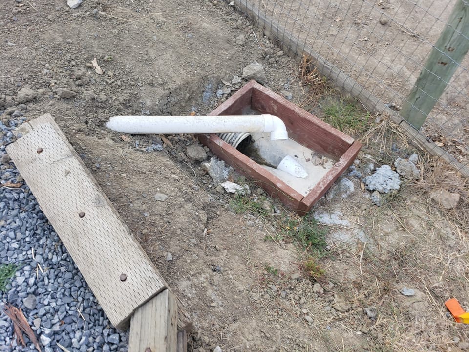 White PVC pipe discharging into a small wooden framed ground basin next to a wire fence.