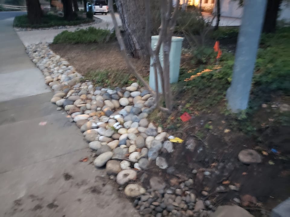 A rock lined dry drainage swale with smooth rounded cobbles running along a concrete driveway.