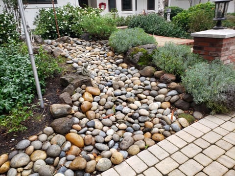 A dry creek bed lined with rounded river rock and boulders curves through a garden beside a paver path.
