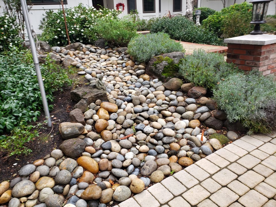 A dry creek bed lined with rounded river rock and boulders curves through a garden beside a paver path.