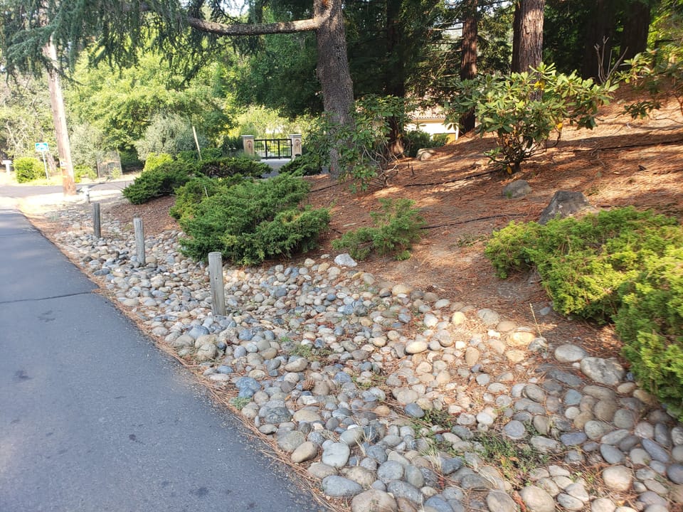 Cobblestone rock drainage swale along the edge of an asphalt road next to a landscaped yard.