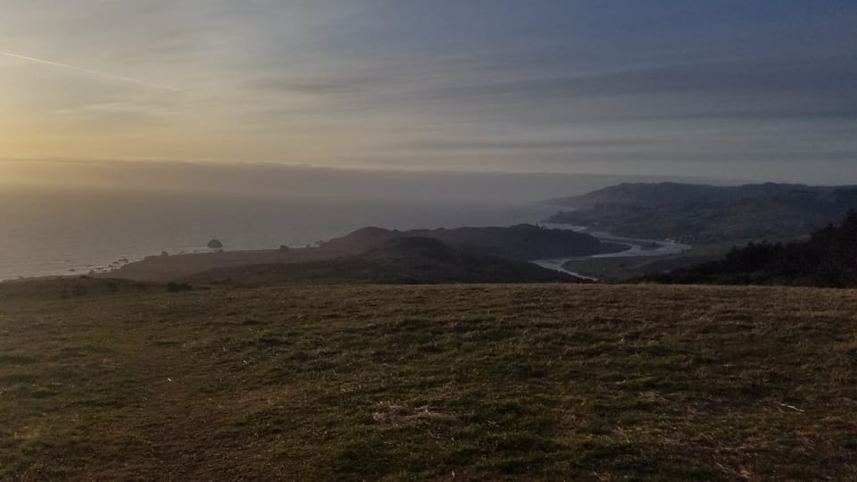 Wide view from a grassy hill overlooking a winding river valley and the Pacific Ocean at dusk.