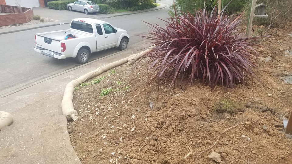 Straw wattles placed along a concrete sidewalk below loose excavated dirt on a sloped residential yard.