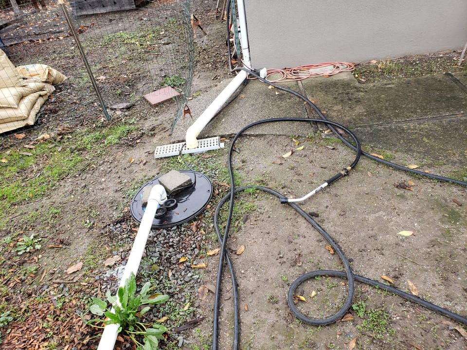 Exterior sump pump basin with PVC pipe, downspout drain, garden hoses, and sandbags beside a gray building wall.