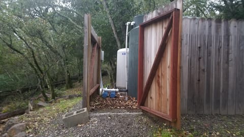 Open wooden gate revealing a green water storage tank, pressure tank, and pump system in a wooded yard.