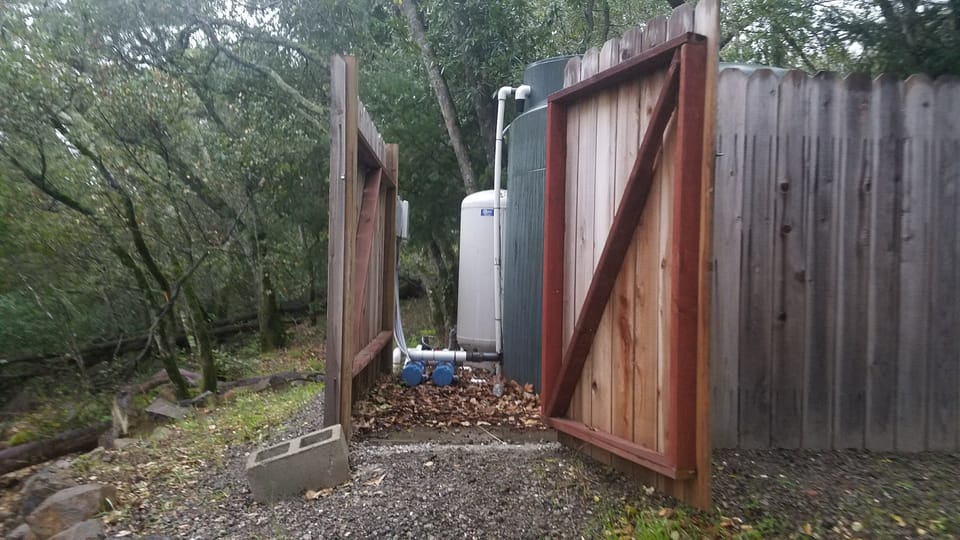 Open wooden gate revealing a green water storage tank, pressure tank, and pump system in a wooded yard.